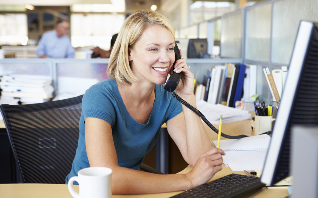 Smiling blonde woman in office holding pencil while using VoIP phone systems and computer